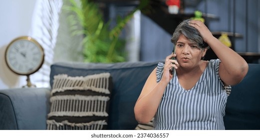 Upset Indian old grey-haired woman sitting on sofa talking on mobile phone answer unpleasant call received bad shocking news. Disappointed worried senior grandmother feel sadness alone at indoor home - Powered by Shutterstock