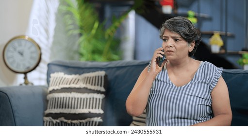 Upset Indian old grey-haired woman sitting on sofa couch talking on mobile phone answer unpleasant call received bad shocking news. Disappointed worried senior grandmother feel sad alone indoor home - Powered by Shutterstock