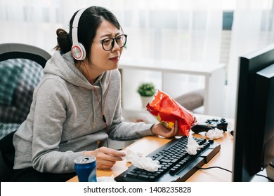 Upset lazy asian woman nerd crying looking at pc computer screen watching sad movie with headphone. unhappy girl geek eat snack chip junk food in hand wipes away tears with used tissue on table.