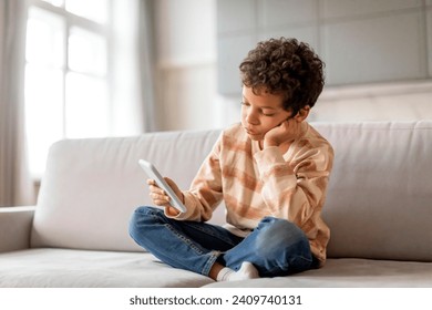 Upset Little Black Boy Using Mobile Phone While Relaxing On Couch At Home, Bored Preteen Male Child Playing Games Or Browsing Internet On Smartphone, Sad Kid Checking New App, Copy Space - Powered by Shutterstock