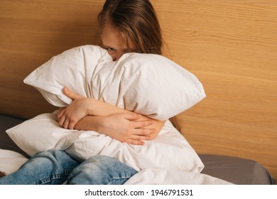 Upset Little Girl Crying Sitting Alone In Fetal Position Hiding Face And Rocking On Bed Hugging Big Pillow And Looking At Camera . Concept Of Child Depression.