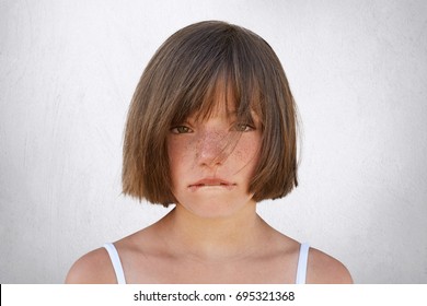 Upset Little Girl Having Quarrel With Her Parents, Looking Innocently Into Camera While Curving Her Lips Isolated Over White Background. Sorrorful Female Child Going To Cry, Biting Her Lower Lip