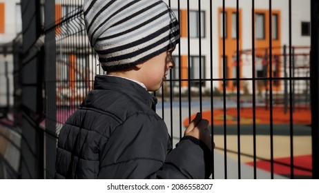 Upset lonely boy looking through metal fence on children palyground and modern school