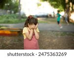 Upset lonely bullied little kid girl cries covering his eyes with his hands feels abandoned abused, sad alone preschool child in kindergarten or playground.