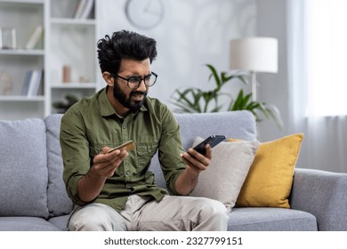 Upset man at home cheated and denied money transfer, hispanic sitting on sofa at home with phone and bank credit card, cheated and disappointed in living room.
