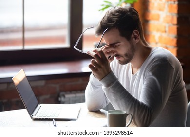 Upset man massaging nose bridge, taking off glasses, feeling eye strain after long work with laptop, sitting in cafe, tired male feeling discomfort after long wearing glasses, bad eye vision concept
