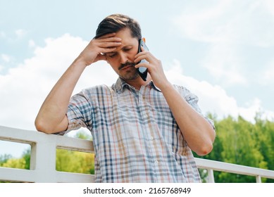 Upset Man With Negative Expression Listening To Bad News On Mobile Phone Outdoors. Sad Guy Holding His Head During An Unpleasant Conversation On Smartphone.