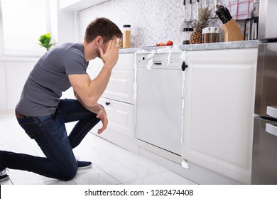 Upset Man Sitting In Front Of Damaged Dishwasher With Foam Coming From It