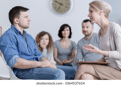 Upset Man Sitting In Group Therapy And Listening To The Psychologist With Concerned Family In The Background