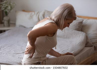 Upset mature woman suffering from backache after sleep, rubbing stiff muscles, unhappy older female sitting on bed at home, feeling discomfort, because of bad posture or uncomfortable bed