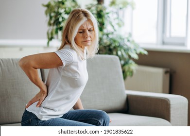 Upset mature woman suffering from backache, unhappy senior blonde sitting on a sofa at living room, feeling discomfort because of pain in back.