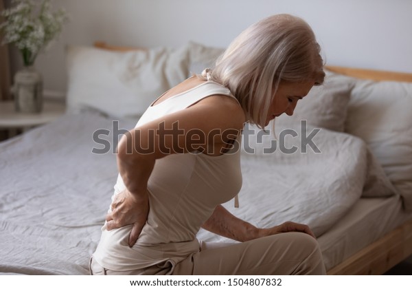 Upset mature woman suffering from backache after\
sleep, rubbing stiff muscles, unhappy older female sitting on bed\
at home, feeling discomfort, because of bad posture or\
uncomfortable bed
