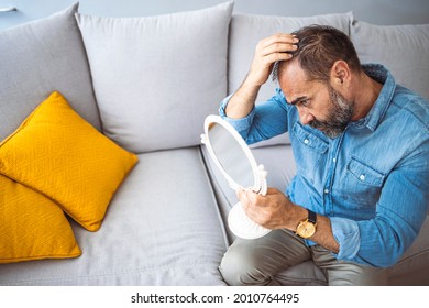Upset middle aged man with alopecia looking at mirror, hair loss concept. Mature men is worried about hair loss. Man controls hair loss and unhappy looking in the mirror