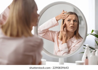 Upset middle aged woman checking wrinkles around her eyes while making beauty routine at home, beautiful mature lady looking in mirror and touching face, feeling tired and stressed, selective focus - Powered by Shutterstock