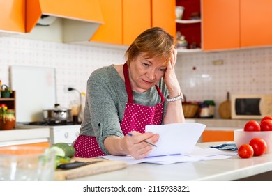 Upset Middle Aged Woman Sitting In Home Kitchen, Reading Bad News In Paper Letter