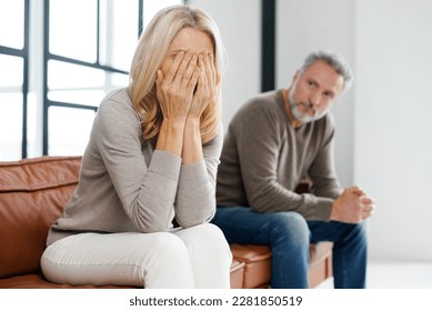 Upset middle-aged couple is arguing at home, frustrated blonde woman covered face with palms and crying, sadness grey-haired mature man on background. Spouses have difficulties, relationship crisis