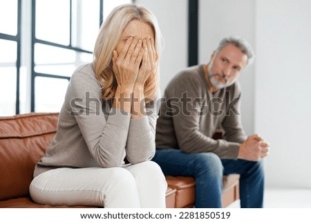 Upset middle-aged couple is arguing at home, frustrated blonde woman covered face with palms and crying, sadness grey-haired mature man on background. Spouses have difficulties, relationship crisis