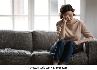 Upset middle-aged woman sit on couch lost in thoughts feel lonely disappointed alone at home, unhappy anxious mature female missing or mourning, senior lady suffer from mental psychological problems