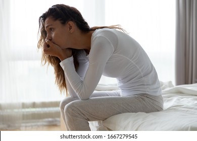 Upset millennial girl sit on bed in bedroom lost in thoughts thinking pondering of problem solution, depressed unhappy young woman look in distance suffer from depression, having personal problems