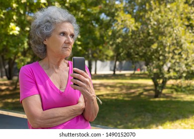 Upset Old Lady Holding Smartphone And Looking Away In Park. Senior Grey Haired Woman In Casual Standing On Walkway And Using Mobile Phone. Bad News Concept