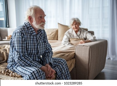 Upset Old Man Sitting On Bed With Sad Look. His Woman Sitting In Bed Under The Blanket And Looking At Him With Sympathy