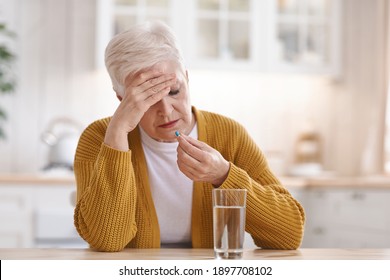 Upset Old Woman Suffering From Headache, Touching Her Forehead And Taking Pill, Kitchen Interior, Copy Space. Grey-haired Elderly Lady Feeling Bad, Having Migraine Or Tension Problem