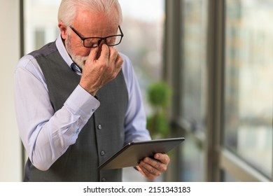 Upset Older Caucasian Man Reading Business Reports On His Tablet Standing In The Office Having A Headache. Depressed Older Businessman Worried About Bad News From The Market