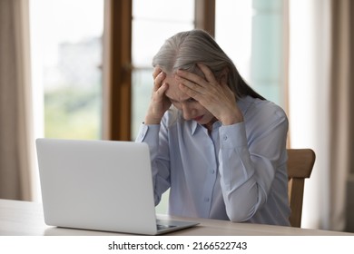 Upset Older Woman Sit At Desk Looks At Laptop Hold Head, Feels Desperate Due To Difficult Task, Modern Tech, Awful News, Bankruptcy Or Crisis, Information Overload. Overwork And Stress At Work Concept