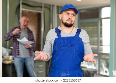 Upset perplexed young bearded builder in blue overalls standing in building under renovation on background of displeased screaming male customer