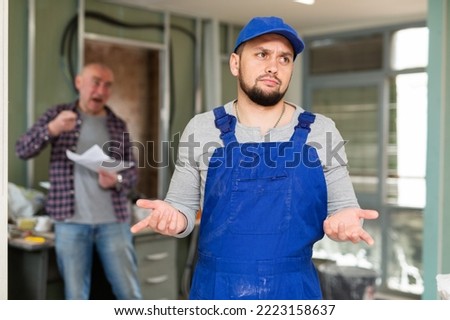 Upset perplexed young bearded builder in blue overalls standing in building under renovation on background of displeased screaming male customer
