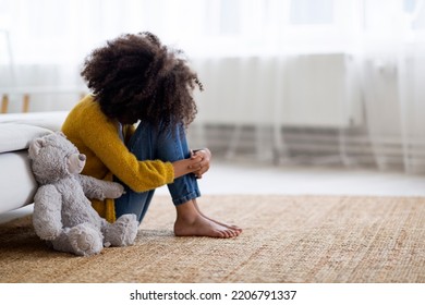 Upset preteen black girl with curly bushy hair sitting on floor with grey teddy bear, crying. African american child feeling lonely at home, missing parents or friends, side view, copy space - Powered by Shutterstock