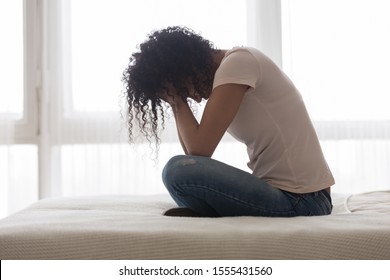 Upset sad African American woman suffering from loneliness side view, sitting in bed alone, holding head in hands, upset girl has psychological troubles, trauma, thinking about problem in bedroom