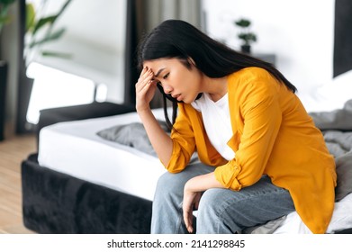 Upset Sad Chinese Brunette Young Woman, In Casual Clothes, Sitting On The Bed At Home, Saddened By Personal Problems, News, Loneliness, Experiencing Stress, Uncertainty