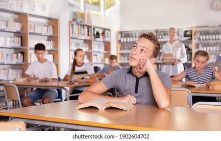 Upset Schoolboy Sitting At Table With Book In The Classroom