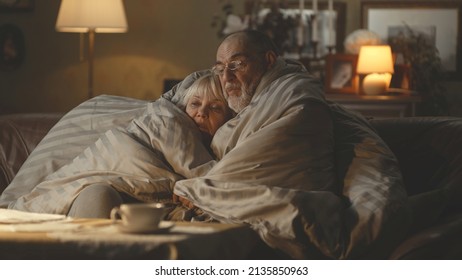 Upset Senior Husband And Wife Embracing Each Other And Speaking Under Warm Duvet While Sitting On Sofa In Cold Apartment During Gas Crisis