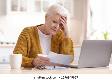 Upset senior lady holdign her head while reading bills, having financial troubles during quarantine. Sad elderly woman sitting in kitchen in front of laptop and holding documents, copy space