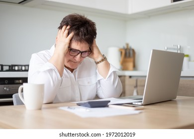 Upset Senior Lady Holdign Her Head While Reading Bills, Having Financial Troubles During Quarantine. Sad Elderly Woman Sitting In Kitchen In Front Of Laptop And Holding Documents