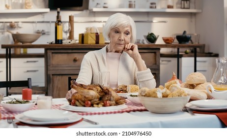 Upset Senior Woman Sitting Alone Near Thanksgiving Dinner On Table In Kitchen