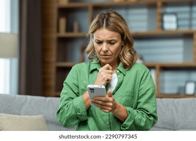 Upset senior woman sitting on sofa at home and holding phone. She looks worriedly at the screen, received a message, bad news. - Powered by Shutterstock