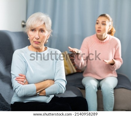 Upset senior woman sitting on sofa in cozy living room while agitated adult daughter reprimanding her. Generation conflicts concept.