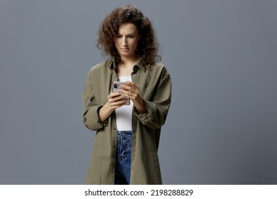 Upset Shocked Confused Curly Beautiful Woman In Casual Khaki Green Shirt Receiving Bad News Using Phone Posing Isolated On Over Gray Blue Background. Social Media Influencer Concept. Copy Space