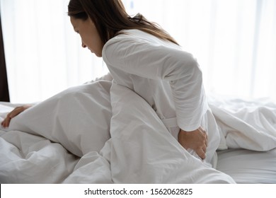 Upset sick young woman wear pajamas touching aching back feel backache pain morning discomfort low lumbar problem sit on bed after bad sleep waking up on uncomfortable mattress, backpain concept