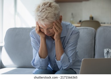 Upset Stressed Older Woman Holds Head With Hands Sit On Sofa Near Laptop, Suffers From Strong Severe Headache, Chronic Migraine After Device Usage, Information Overload, Awful News, Bank Debt Concept