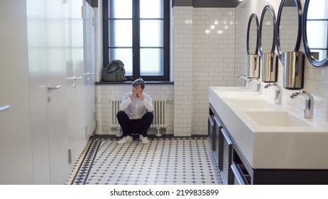Upset teen schoolboy sitting on floor in school toilet alone. Frustrated and lonely teenager sitting on floor in campus bathroom