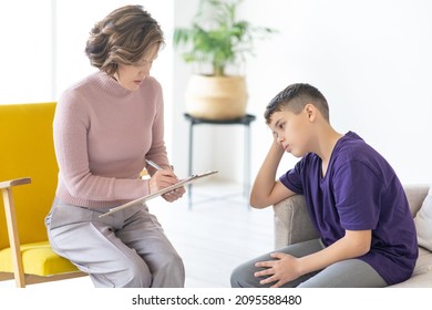 Upset teenager holding his head. Woman psychologist writes on clipboard. Appointment with teenage psychologist. School psychologist.