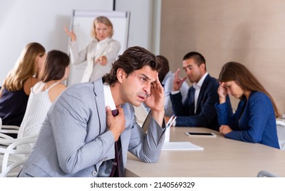 Upset And Tired Businessman Sitting At Desk In Office On Background With Angry Female Boss Scolding Subordinates