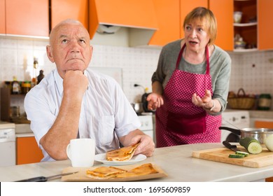 Upset And Tired Senior Man Sitting At Home Table During Lunch On Background With Disgruntled Wife Scolding Him