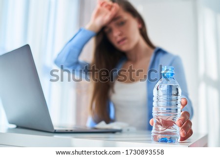 Upset tired working woman suffering from heat, thirst and hot weather cools down with cold water bottle during online work at computer at warm summer day