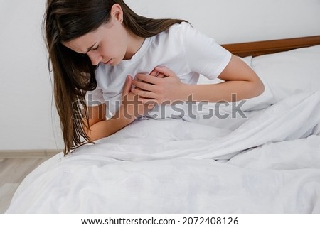 Upset tired young brunette female feeling pain ache touching chest having heart attack, sad unhappy worried girl suffers from heartache sit on bed at home, infarction or woman heart disease concept