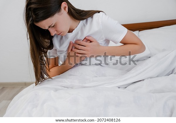 Upset tired young brunette female feeling pain\
ache touching chest having heart attack, sad unhappy worried girl\
suffers from heartache sit on bed at home, infarction or woman\
heart disease concept